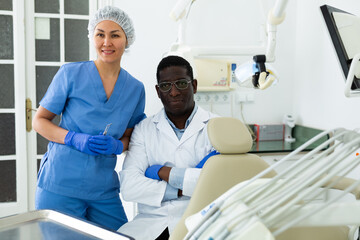 Positive confident african american dentist sitting with asian female assistant in modern dental clinic office ..