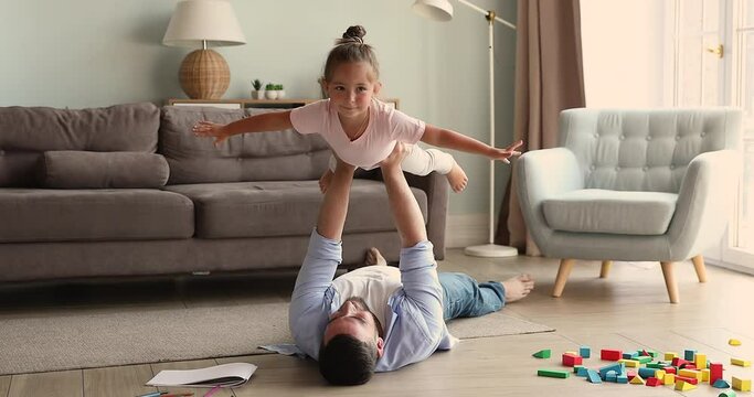 Young dad lift on stretched strong arms cute little daughter, lying on warm floor in cozy living room, spend weekend leisure at modern home. Daydreams about family travel, funny active games concept