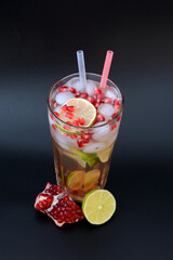 Pomegranate juice with lime in a tall glass with ice and straws on a black background.