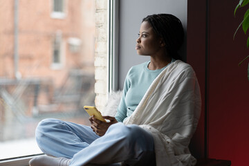 Focused African American woman covered with blanket sitting on windowsill, holding a phone, thinking about something. Serious millennial female deep in thoughts looking out window, waiting for message - Powered by Adobe