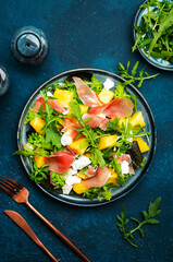 Delicious summer fresh salad with cantaloupe melon, prosciutto, soft cheese and arugula on blue table background, top view, copy space