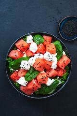 Summer Watermelon juicy salad bowl with feta cheese, spinach and black sesame seed, dark table background, top view, negative space