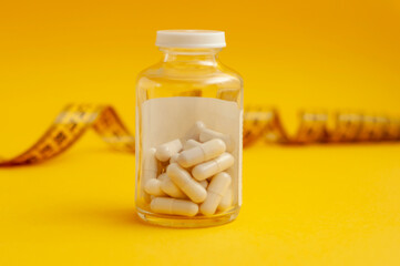 glass jar with tablets in capsules stands in front of a measuring tape on a yellow background, a horizontal snapshot. The concept of weight loss, fat burner, vitamins, sports nutrition. © Lyubov