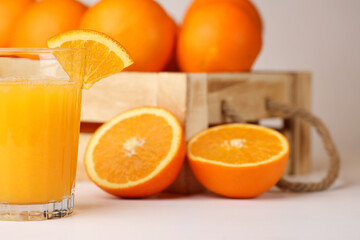 A glass of fresh orange juice, a slice of an orange on it, wooden box with rope handles full of oranges, one sliced orange on the side