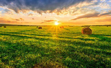 Scenic view at picturesque burning sunset in a green shiny field with hay stacks, bright cloudy sky , trees and golden sun rays, summer valley landscape