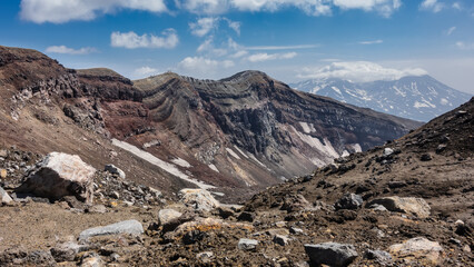 At the top of an active volcano, a layered structure of steep slopes, rocky soil is visible. There are tiny silhouettes of people on the edge of the crater. Blue sky, clouds. Kamchatka. Gorely Volcano