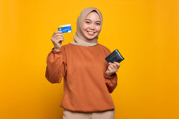 Smiling young Asian woman in orange shirt holding wallet and showing credit card isolated over yellow background