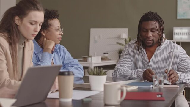 Waist up slowmo of African American engineer presenting windmill model to diverse colleagues, sitting together at office desk developing project on alternative energy sources