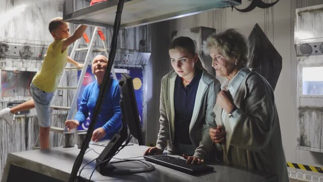 Grandmother and granddaughter uisng computer in escape room to solve conundrum. Grandfather and grandson standing in background and looking at map.
