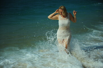 A woman of 30-40 years old in a white lace dress stands on the ocean, and waves of clear water reach her feet with a sea of different colors from turquoise to dark. High quality photo
