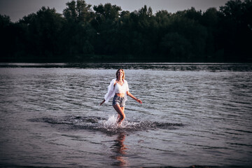 A young woman runs in the water. A smiling blonde in a white top, shirt, denim shorts runs along the river against the background of a beautiful landscape. 