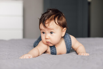 Naklejka premium Portrait of pretty baby wearing denim romper lying on gray blanket at home. Looking away infant child crawl on bed in bedroom, explore world. Selective focus, free copy space. Childcare concept.