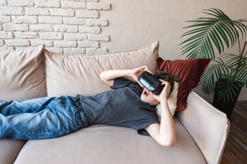 teenage boy in virtual reality glasses lies at home on the couch and screams and gesticulates in...
