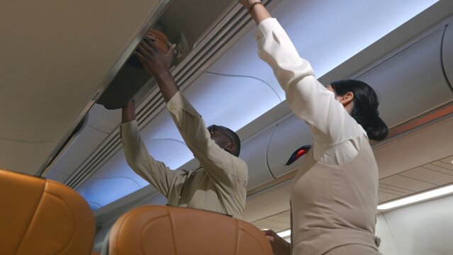 Friendly Flight Attendant. Air Hostage Helping Passenger Businessman Put Luggage On Top Shelf Or Cabin Compartment On Airplane, Woman Help Black Man Lift Suitcases Putting Overhead Locker On Airplane