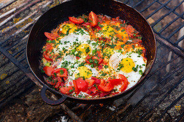 Eggs prepared with vegetables on a frying pan on the fire in the forest. Cooking scrambled eggs over an open fire. Breakfast in nature. Tourist food.