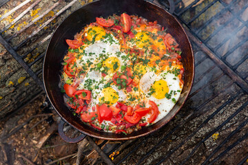 Eggs prepared with vegetables on a frying pan on the fire in the forest. Cooking scrambled eggs over an open fire. Breakfast in nature. Tourist food.