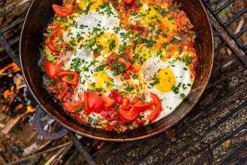 Eggs prepared with vegetables on a frying pan on the fire in the forest. Cooking scrambled eggs over an open fire. Breakfast in nature. Tourist food.