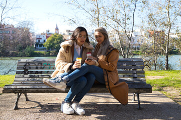 A lesbian couple sitting on a bench checking their social networks over a cup of coffee. The women are young and recently married. Concept lgtb, gays and lesbians. Rights and equality