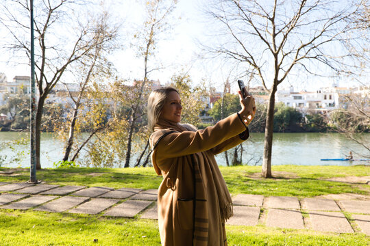Young, blonde, beautiful, lesbian woman taking a photo with her mobile phone in the park. In the background you can see the river and a house. Concept lgtb, gay, equality.