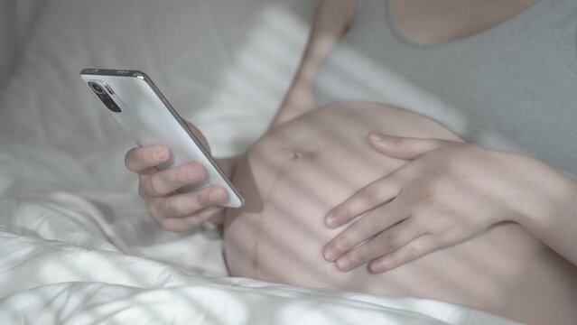 A pregnant woman uses the phone and strokes her belly while relaxing in a home bed. Using a smartphone before the birth of a child. Women's health, remote consultation with a doctor, pregnancy period