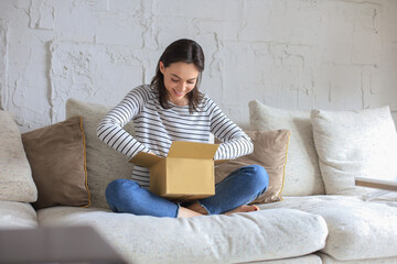 Beautiful young woman is holding cardboard box and unpacking it sitting on sofa at home. - obrazy, fototapety, plakaty