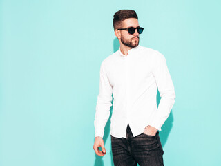 Portrait of handsome confident model. Sexy stylish man dressed in white shirt and jeans. Fashion hipster male posing near blue wall in studio. Isolated. In sunglasses