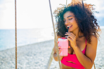Young beautiful woman with pink milk shake on the beach chilling on swing alone