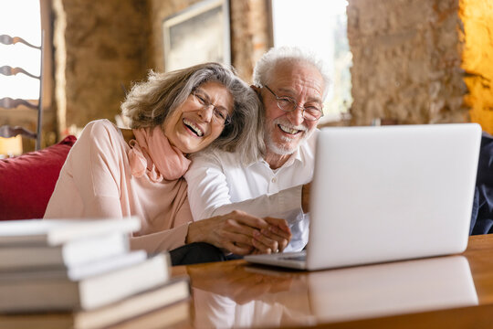 Happy Senior Couple Sharing Laptop Sitting On Sofa In Boutique Hotel