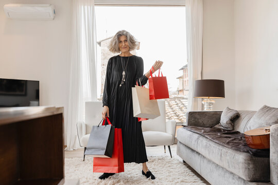 Smiling senior woman with sopping bags standing by sofa at boutique hotel