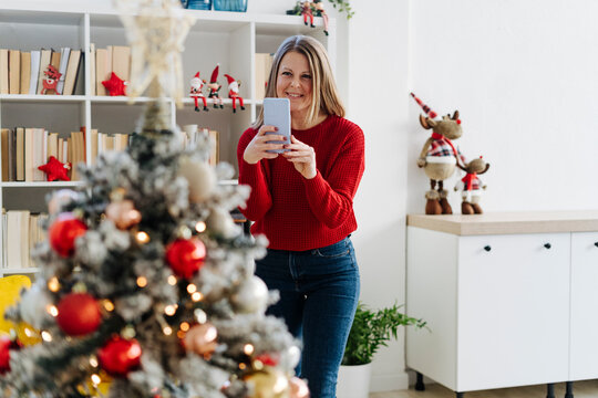 Happy woman photographing decorated Christmas tree through mobile phone at home