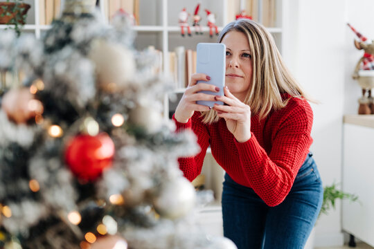 Woman photographing Christmas tree through mobile phone at home