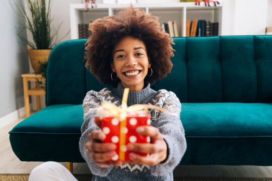 Happy young woman giving Christmas present sitting by sofa at home