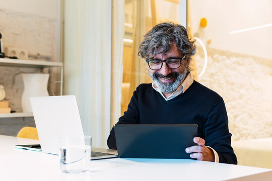 Happy senior freelancer using tablet PC sitting with laptop at table