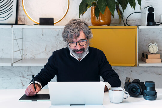 Senior freelancer with digitized pen writing on tablet PC sitting with laptop at table