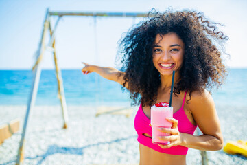 latin brazilian woman with curly long hairstyle and pink bikini drinking strawberry smoothie in beach on the swing