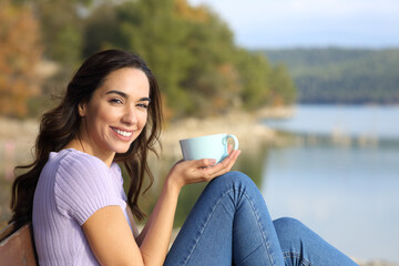 Happy woman holding coffee mug looks at camera in nature