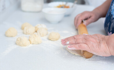 Senior woman hands rolling out the dough with a rolling pin on a white kitchen table with blurred grated apple and sugar on background. Selective focus. Process of making pies with apple filling