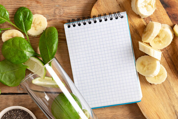 Notebook with ingredients for cooking detox smoothie. Glass with spinach, banana, lime and chia seeds on wooden table top view. Healthy food concept