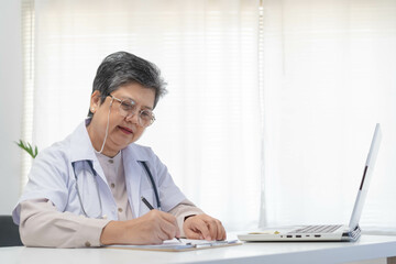 Naklejka na ściany i meble Senior asian physician, general woman doctor wearing glasses and white uniform, mature female with stethoscope, working on laptop computer, sitting in medical office. Healthcare and medical.
