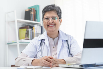 Fototapeta premium Smile senior asian physician, general woman doctor wearing glasses and white uniform, mature female with stethoscope, working on laptop computer, sitting in medical office. Healthcare and medical.