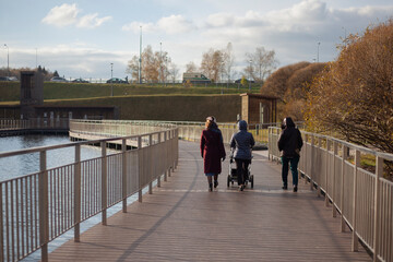 Family is walking in park. Three women walk across bridge over water. People in city in fall. Details of park architecture.