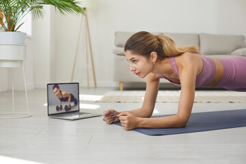 Determined active young woman doing sports workout on floor mat at home, watching online video,...