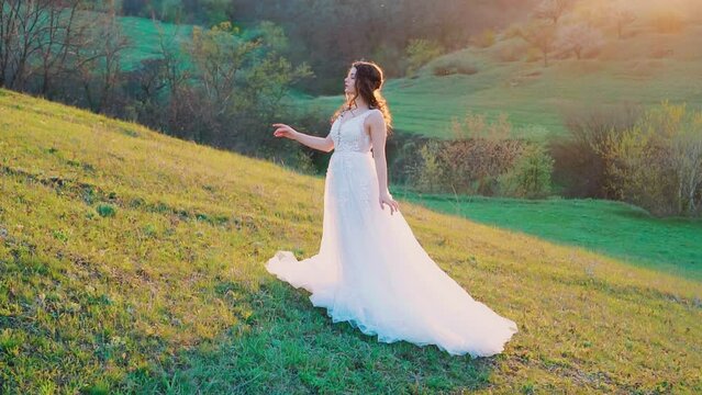 Woman bride in white luxurious puffy wedding dress, creative hairstyle design. Adult girl standing on green fresh grass lawn, spring meadow hill, sunset sun rays. Ball gown long train. Summer nature
