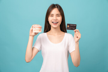 Smiling Asian woman wearing braces showing pointing finger and holding tooth model on blue background.