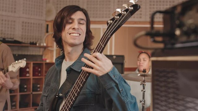 Medium slowmo of young emotional rock band of three playing musical instruments at professional music studio while recording new song
