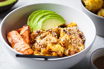 Vegan lunch bowl with baked cauliflower, sweet potato, avocado and peanut butter dressing. Healthy food concept.