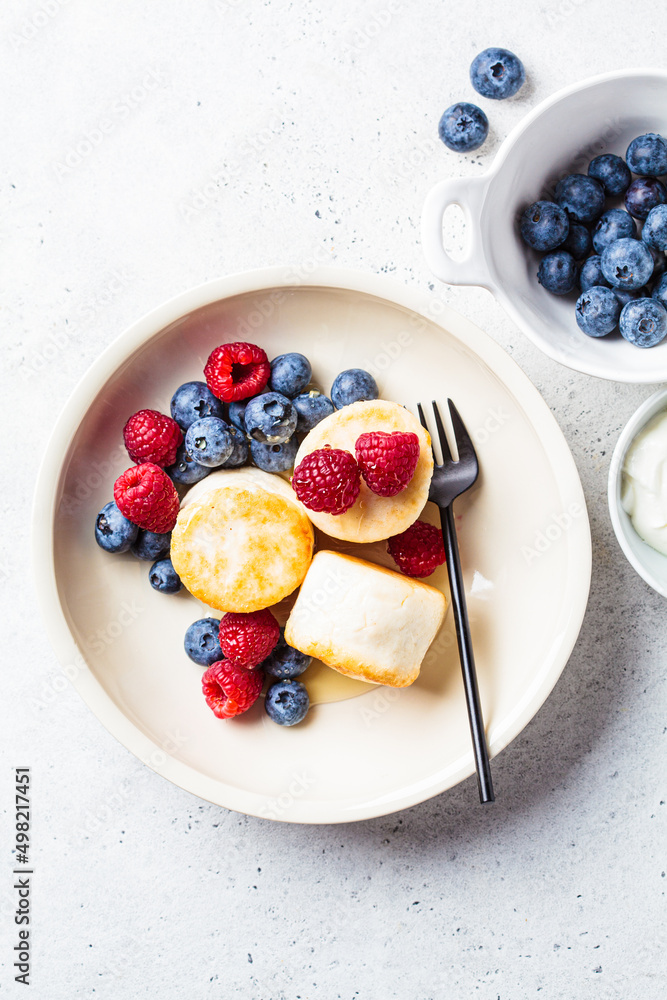 Poster Cottage cheese pancakes with raspberries, blueberries and syrup in white bowl, gray background. Russian cuisine concept.