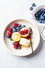 Cottage cheese pancakes with raspberries, blueberries and syrup in white bowl, gray background. Russian cuisine concept.