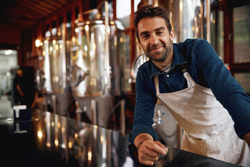 He will pour you anything you want. Portrait of a cheerful young barman leaning on the bar counter waiting for customers to serve inside of a beer brewery.