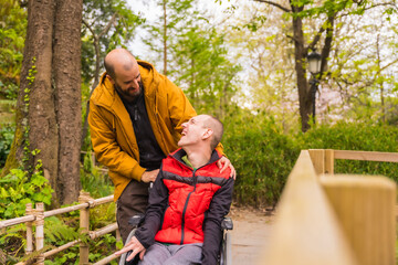 Fototapeta premium Paralyzed young man in the wheelchair being pushed by a friend in a public city park, looking at each other and smiling